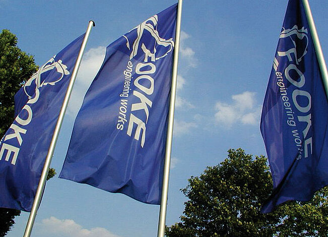 Drei blaue Fahnen mit dem FOOKE-Logo wehen im Wind.